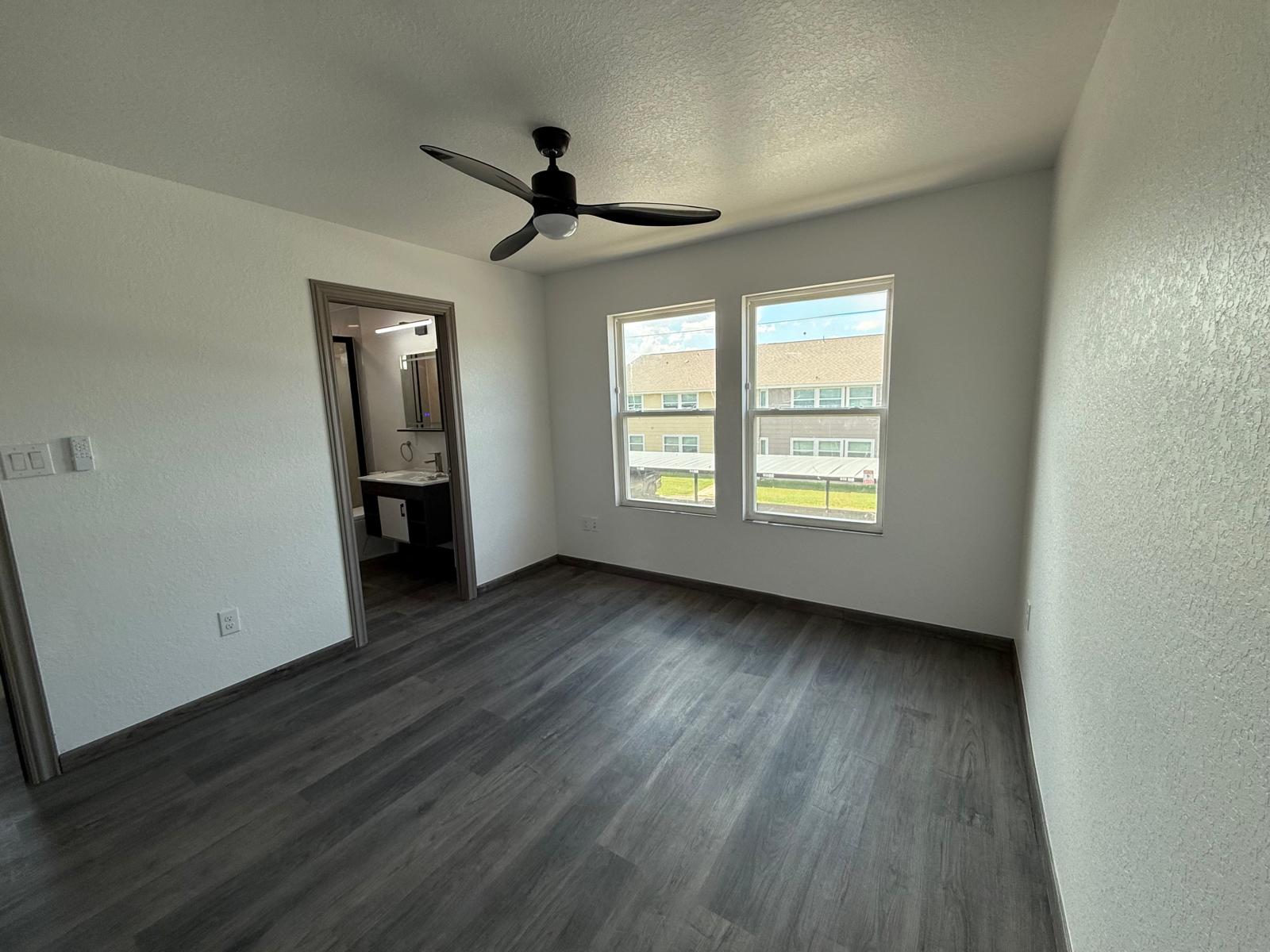 Empty apartment bedroom with two windows, gray flooring, and ceiling fan